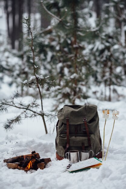 冬のキャンプ。旅行のリュックサック、地図、雪の上に2つのマグカップとマシュマロと一緒にボンファイア