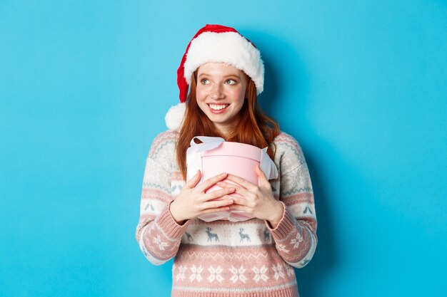 冬とお祝いのコンセプト。彼女のクリスマスプレゼントを抱き締めて左を見て、幸せそうに笑って、青い背景の上に立っているサンタ帽子の夢のような赤毛の女の子。