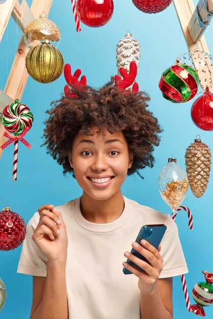 Winter celebration and festive event concept. Smiling glad dark skinned woman receives greeting sms on smartphone during New Year eve wears reindeer hoop prepares for winter holidays. Cozy atmosphere