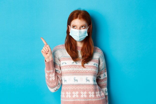 Winter, covid-19 and quarantine concept. Image of cute redhead girl in face mask and sweater, pointing upper left corner, picking product, standing over blue background