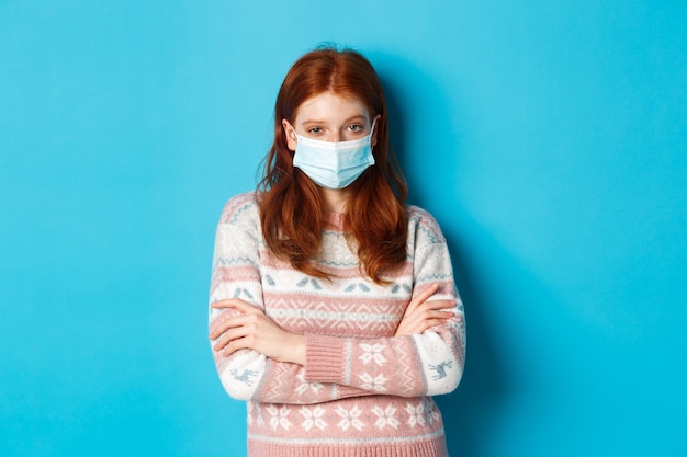 Winter, covid-19 and quarantine concept. Skeptical redhead girl in sweater and medical mask, cross arms on chest and staring with disbelief, standing over blue background