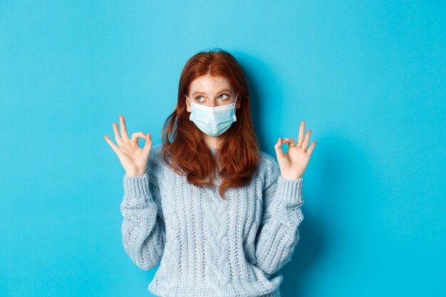 Winter, covid-19 and social distancing concept. Satisfied young redhead woman in face mask showing alright, okay gestures and looking left at promo, blue background.