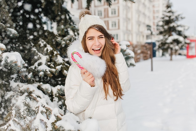 Winter frozen time of funny amazing woman having fun with pink lollypop on street. Young joyful woman enjoying snowing in warm jacket, knitted hat, expressing positivity. Delicious, sweet winter time.