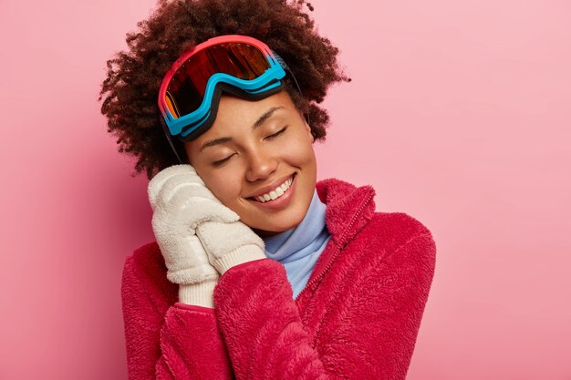 Winter fun, recreation and lifestyle concept. Dark skinned woman wears snowboard glasses leans at both hands in white gloves, remembers pleasant moment from vacation