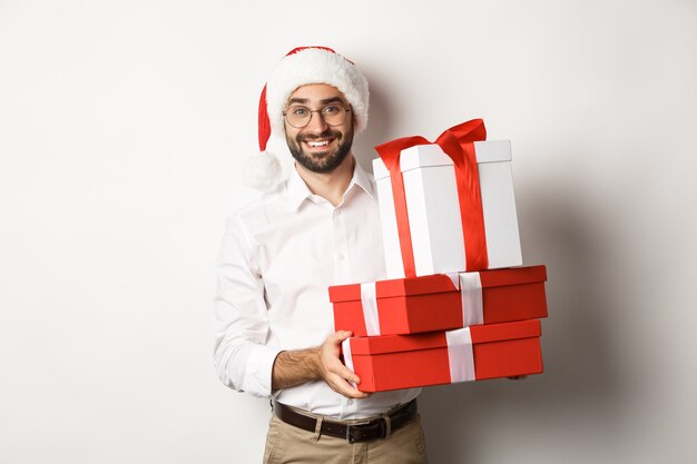 冬の休日とお祝い。幸せな男はクリスマスプレゼントを持ってきて、贈り物を持って、サンタの帽子をかぶって、立っています