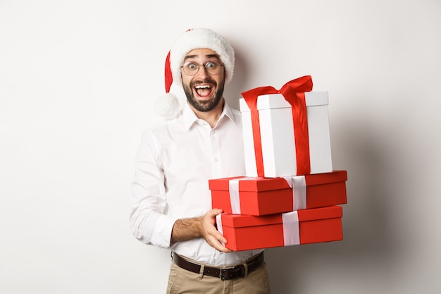 冬の休日とお祝い。幸せな男はクリスマスプレゼントを持ってきて、贈り物を持って、サンタの帽子をかぶって、立っています