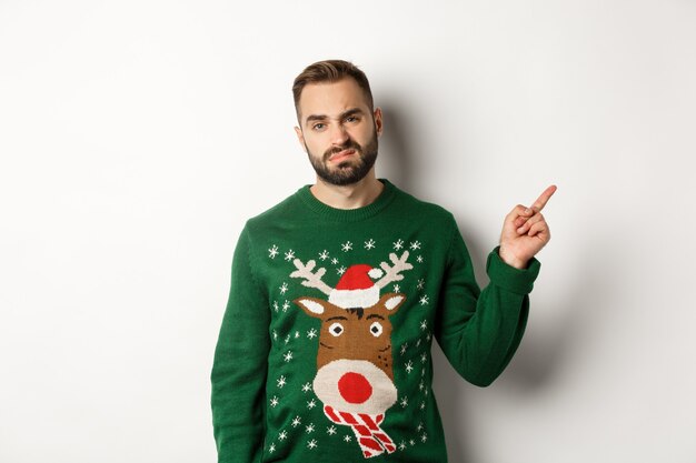 Winter holidays and christmas. Displeased and grumpy guy complaining, wearing funny sweater, pointing finger at upper right corner promo, white background.