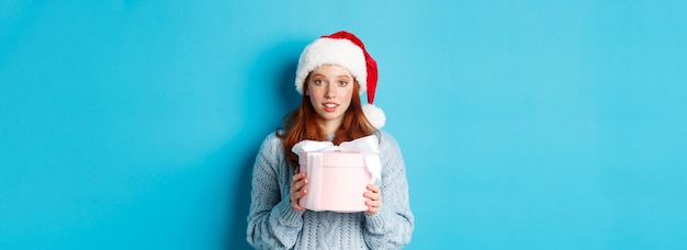 Vacanze invernali e concetto di vigilia di natale ragazza rossa carina che indossa il cappello di babbo natale che tiene il regalo del nuovo anno