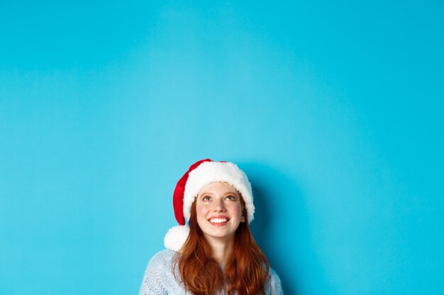 겨울 방학 및 크리스마스 이브 개념입니다. 산타 모자를 쓴 귀여운 빨간 머리 소녀의 머리는 아래에서 나타나 복사 공간을 올려다보고 로고를 쳐다보고 파란색 배경 위에 서 있습니다