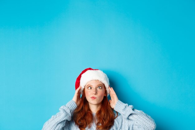 冬の休日とクリスマスイブのコンセプト。サンタの帽子をかぶった面白い赤毛の女の子の頭は、下から目を細めて、愚かな顔をして、青い背景のコピースペースの近くに立っています