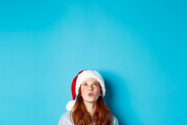 겨울 방학 및 크리스마스 이브 개념입니다. 산타 모자를 쓴 예쁜 빨간 머리 소녀의 머리는 아래에서 나타나 인상적인 로고를 올려다보고 프로모션 제안, 파란색 배경을 보고 있습니다.