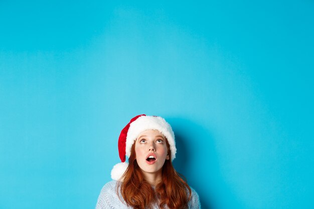 Зимние каникулы и концепция кануна Рождества. Голова красивой рыжей девушки в новогодней шапке появляется снизу и смотрит на логотип, изумленно видя промо-предложение, синий фон.