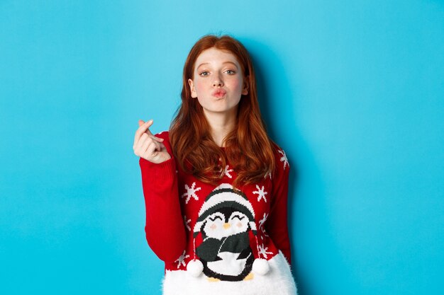 Winter holidays and Christmas Eve concept. Lovely redhead girl in xmas sweater, showing heart sign and pucker lips for kiss, standing over blue background