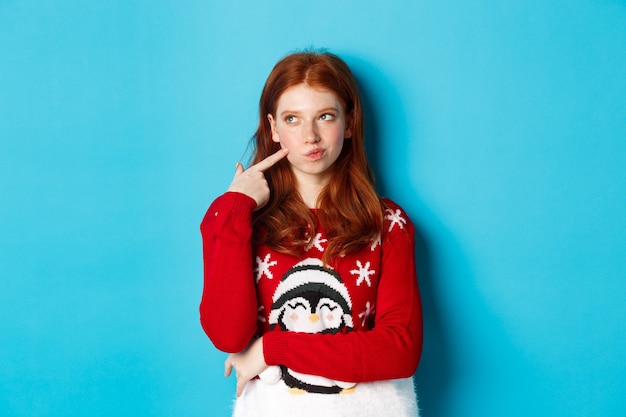 겨울 방학 및 크리스마스 이브 개념입니다. 크리스마스 스웨터를 입은 예쁜 빨간 머리 소녀, 사려깊은 뺨을 만지고 웃고, 선택하고, 왼쪽 상단 모서리를 보고 생각하고, 파란색 배경을 생각합니다.