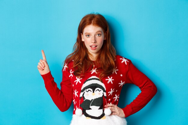 冬の休日とクリスマスイブのコンセプト。クリスマスセーターの生意気な赤毛の女の子、左上隅を指してカメラを見つめ、プロモーションのオファーをほのめかして、青い背景