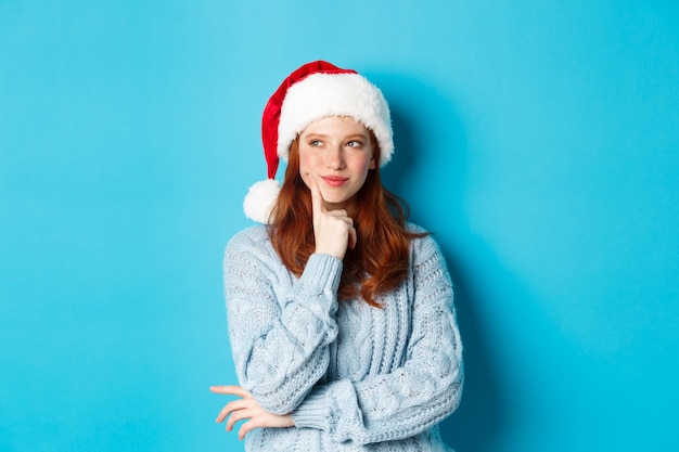 겨울 방학 및 크리스마스 이브 개념입니다. 주근깨가 있는 바보 같은 빨간 머리 소녀, 산타 모자를 쓰고 생각하고, 새해 축하를 계획하고, 파란색 배경 위에 서 있습니다.