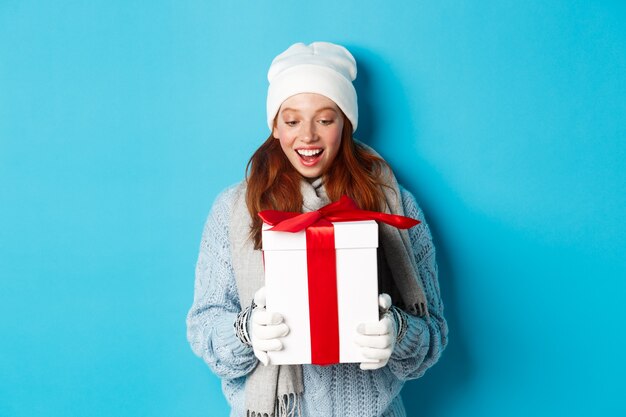 겨울 방학 및 크리스마스 이브 개념입니다. 비니와 스웨터를 입은 놀란 귀여운 빨간 머리 소녀는 새해 선물을 받고 현재 놀란 표정으로 파란 배경 위에 서 있는 모습을 보고 있습니다.