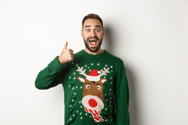 겨울 방학과 크리스마스. 흥분한 백인 남자가 엄지손가락을 치켜들고 놀란 표정을 하고 흰색 배경에 녹색 스웨터를 입고 서 있다