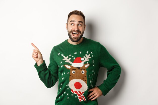 Зимние каникулы и рождество. Счастливый человек в забавном свитере, указывая пальцем на логотип в верхнем левом углу, показывая рекламу, на белом фоне.