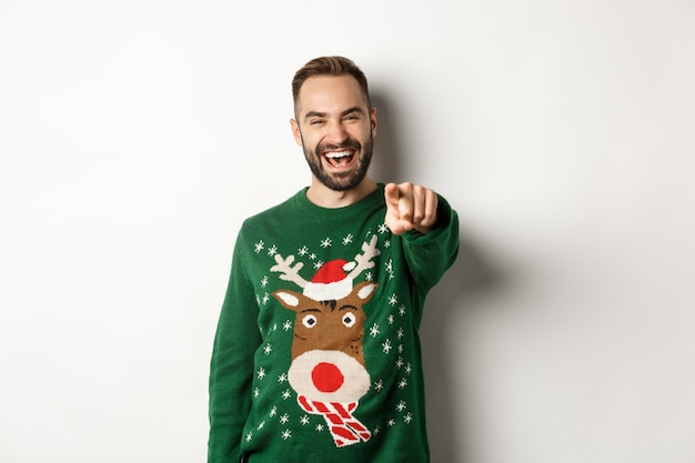 Winter holidays and christmas. Happy young man celebrating New Year eve, pointing finger at you and laughing, standing over white background