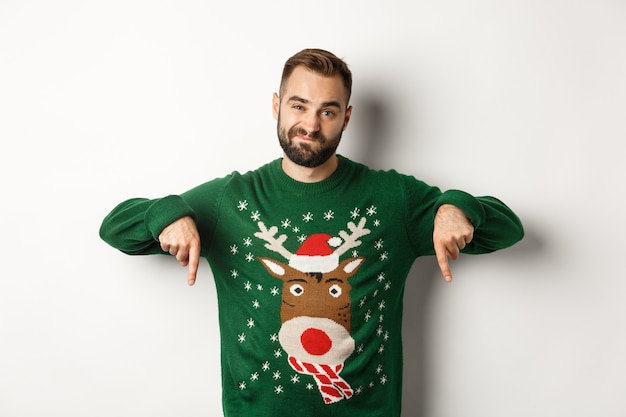 Зимние каникулы и рождество. Скептически настроенный бородатый мужчина выглядит разочарованным, указывая пальцами вниз в знак неприязни к промо, стоя на белом фоне