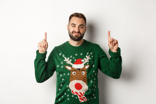 Winter holidays and christmas. Unamused bearded guy in funny sweater pointing fingers up, showing something unpleasant, white background