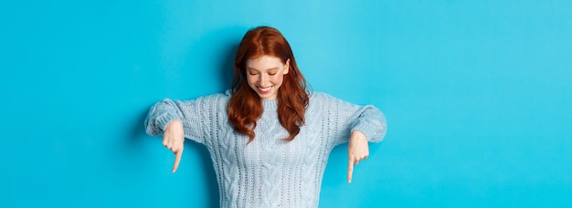 Foto gratuita vacanze invernali e concetto di persone ragazza rossa allegra in maglione che punta le dita verso il basso e guarda