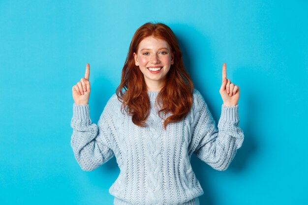 冬の休日と人々の概念。セーターを着た陽気な赤毛の少女が指を上に向け、ロゴバナーを表示し、笑顔で、青い背景の上に立っています。