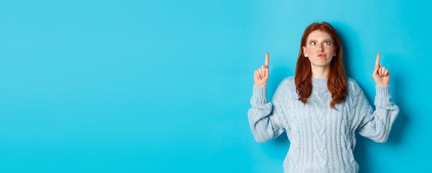 Foto gratuita vacanze invernali e concetto di persone ragazza rossa divertente e sciocca in maglione che punta le dita verso l'alto e s