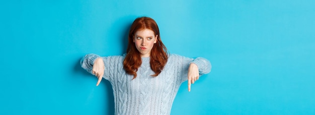 Free photo winter holidays and people concept indecisive redhead girl in sweater pointing fingers down and thin