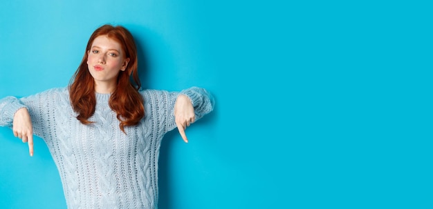 Winter holidays and people concept thoughtful teenage redhead girl in sweater pointing fingers down