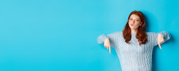 Winter holidays and people concept thoughtful teenage redhead girl in sweater pointing fingers down