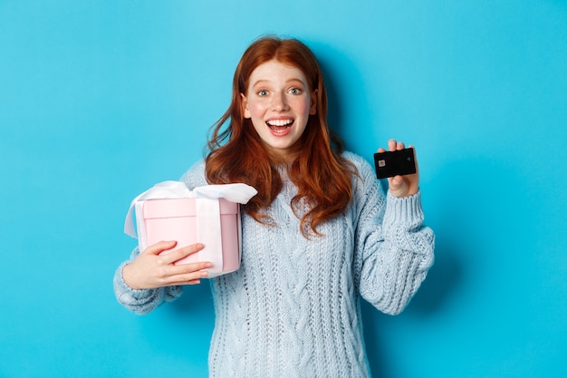 Winter holidays promo offer concept. Cheerful redhead woman holding Christmas gift and credit card, staring at camera amazed, standing over blue background