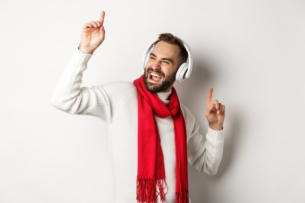 Winter holidays and technology concept. Happy man dancing to music in headphones, standing over white background in sweater