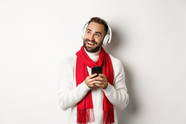 겨울 방학 및 기술 개념입니다. 헤드폰으로 음악 팟캐스트를 듣고 휴대폰을 들고 빈 공간, 흰색 배경을 바라보는 행복한 남자