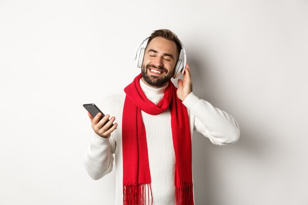 Winter holidays and technology concept. Satisfied man listening music in headphones with closed eyes, smiling with pleasure, holding smartphone, white background.