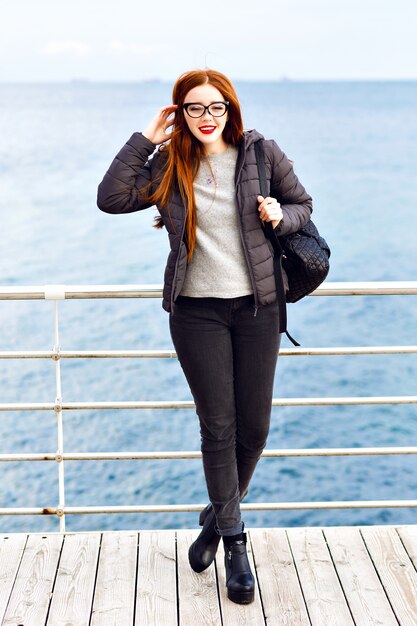 winter lifestyle portrait of pretty hipster ginger woman walking at seaside, total black stylish outfit, cold rainy weather, backpack, leather boots, lonely.