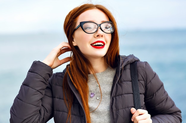 Free photo winter lifestyle portrait of pretty hipster ginger woman walking at seaside, total black stylish outfit, cold rainy weather, backpack, leather boots, lonely.