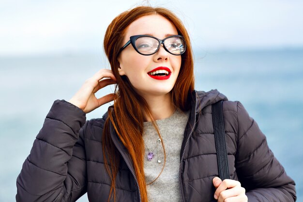 Winter lifestyle portrait of pretty hipster ginger woman walking at seaside, total black stylish outfit, cold rainy weather, backpack, leather boots, lonely.