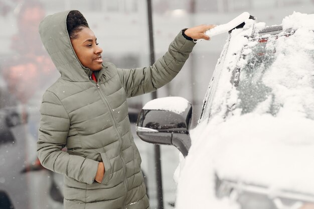 Winter portrait of african woman cleaning snow from a car. Woman in a green jacket.