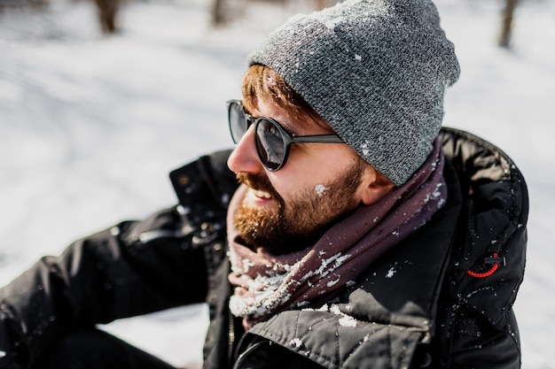 Зимний портрет хипстерского мужчины с бородой в серой шляпе, расслабляющегося в солнечном парке со снежинками на одежде