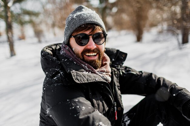 Winter portrait of hipster man with beard in grey hat relaxing in sunny park with snowflakes on clothes