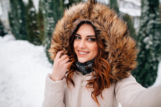 冬のセルフ。毛皮のフードの美しい少女は雪の冬の日にセルフを取る