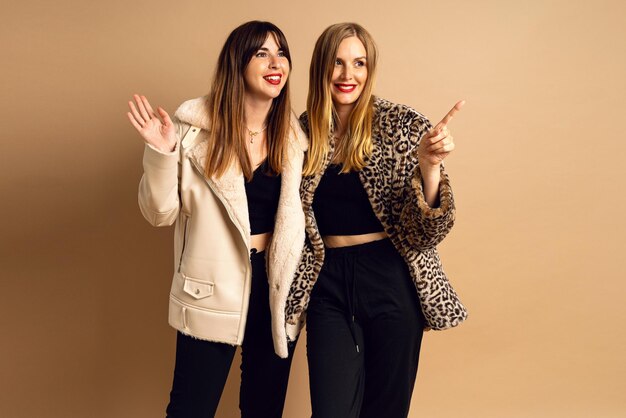 Winter studio portrait of happy two stylish woman wearing warm fur coats posing at beige background glamour style