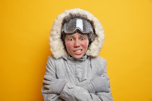 Winter time and cold concept. Freezing woman trembles and keeps hands crossed over body to warm up during blizzard goes skiing in mountains dressed in jacket and ski goggles covered with snow