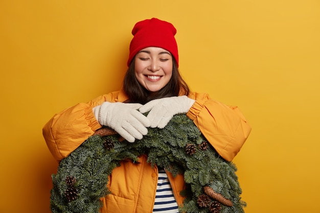 Foto gratuita la donna invernale ha un sorriso a trentadue denti, compra una ghirlanda verde, indossa un cappello rosso, un cappotto giallo e guanti bianchi, si aspetta la vigilia di natale, posa contro il muro giallo.