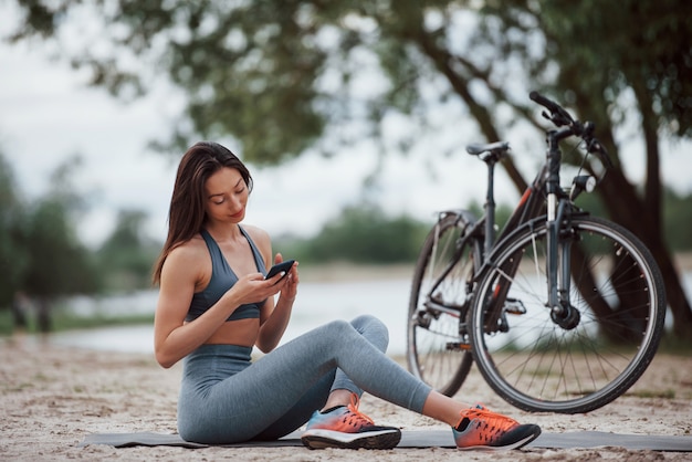 Беспроводные технологии. Велосипедистка с хорошей формой тела сидит возле своего велосипеда на пляже в дневное время