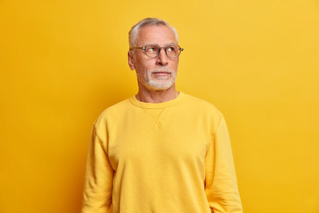 오른쪽에 결정된 사려 깊은 표정으로 집중된 현명한 지적인 수염 난 남자는 두꺼운 회색 수염이 투명한 안경과 노란색 벽 위에 고립 된 캐주얼 점퍼를 착용합니다.