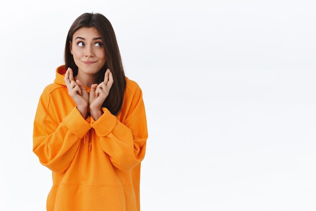 Wishful dreamy gorgeous brunette girl in orange hoodie smiling and looking aside while maing wish cross fingers good luck have something in mind and dream to make it real white background