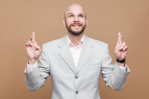 Wishing hopeful portrait of handsome middle aged bald bearded businessman in classic light gray suit standing clenching teeth and crossed fingers. indoor studio shot, isolated on brown background.